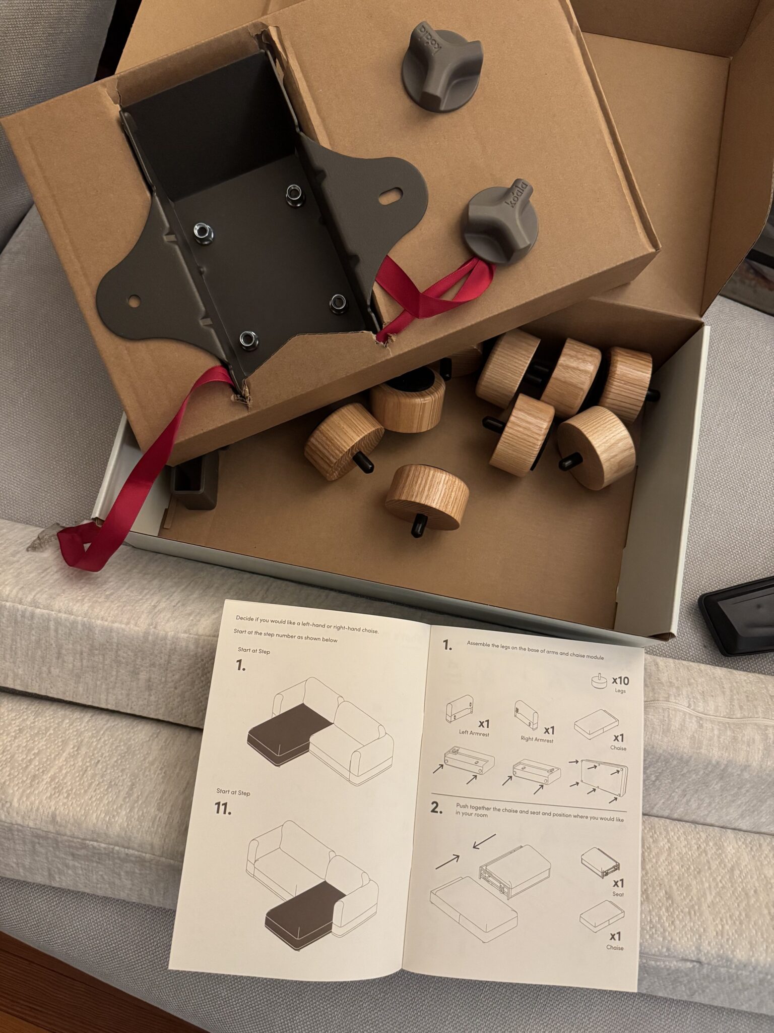An open box with metal parts, wooden knobs, gray plastic pieces, and an instruction manual with assembly diagrams are laid out on a gray couch.