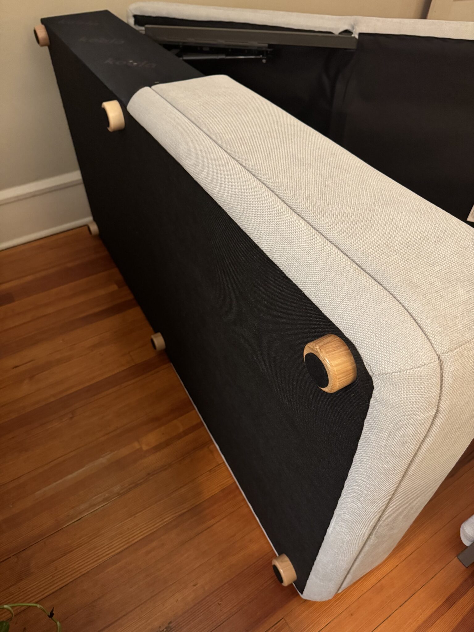 A light gray upholstered couch turned on its side, exposing black fabric on the underside and wooden legs, on a hardwood floor.