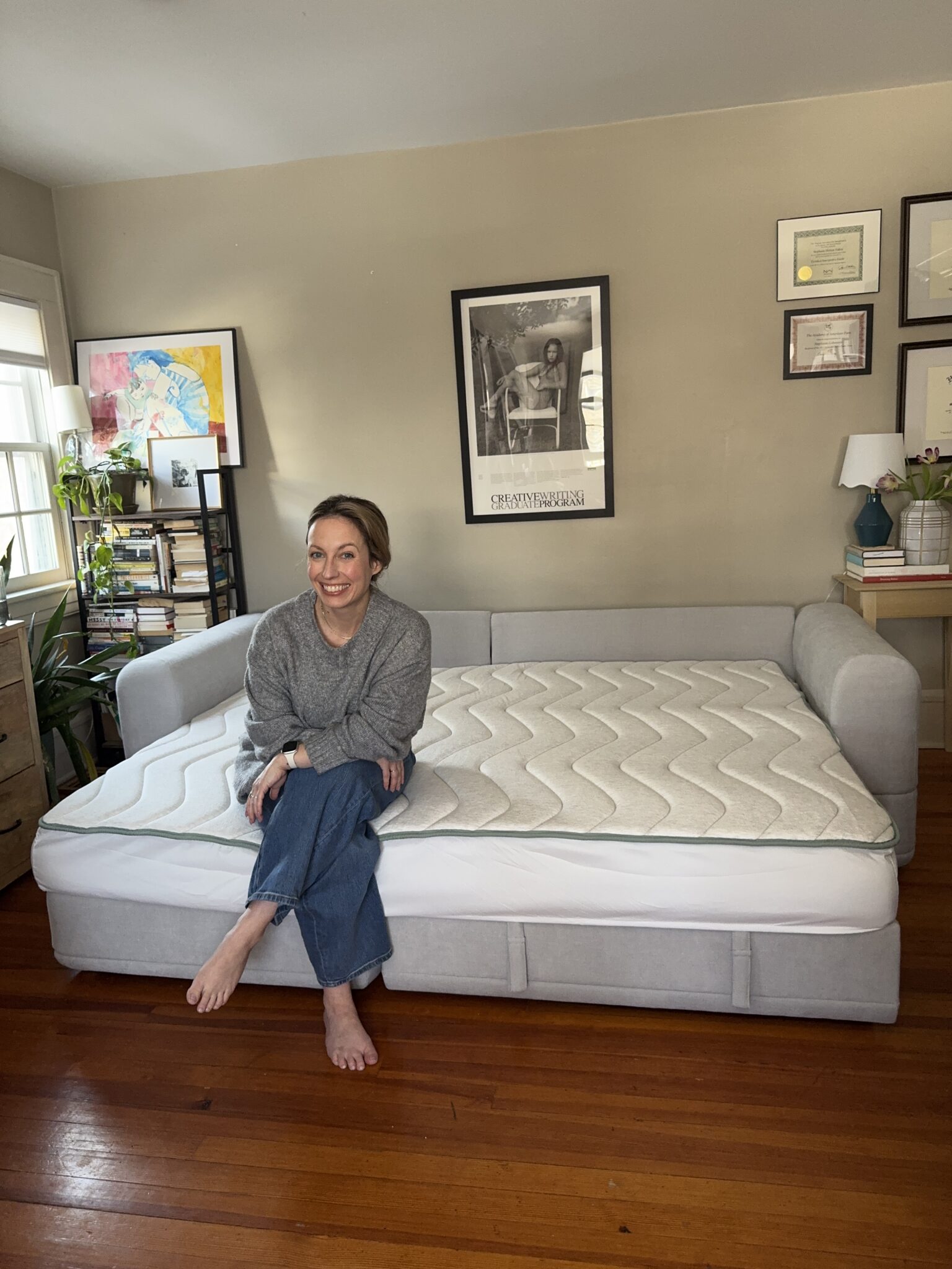 A woman sits on the edge of a large, light gray sofa bed with a mattress topper in a tidy room decorated with framed art and books.