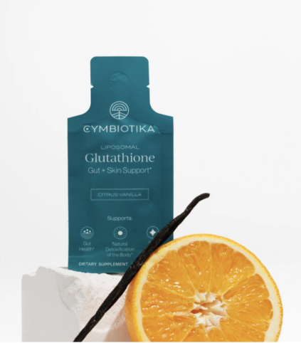 A packet of Cymbiotika Liposomal Glutathione supplement stands upright behind a sliced orange and a vanilla bean on a white surface.