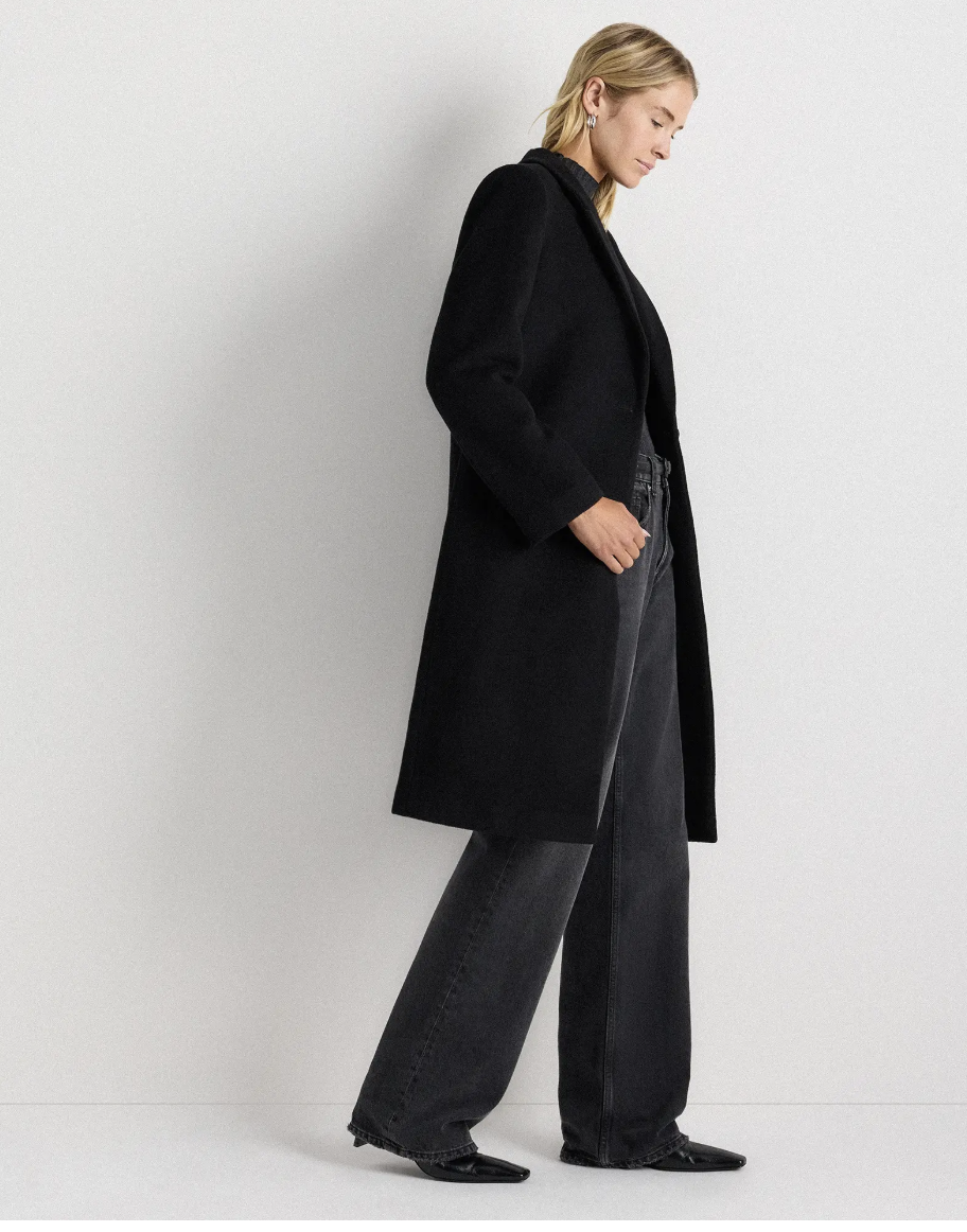 A person with light hair wears a long black coat, dark wide-leg jeans, and black pointed shoes, standing against a plain white wall.