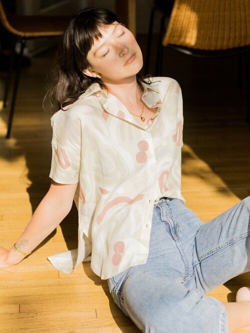 A woman sits on a wooden floor, eyes closed, leaning back with sunlight on her face. She wears a patterned short-sleeve shirt and light blue jeans.