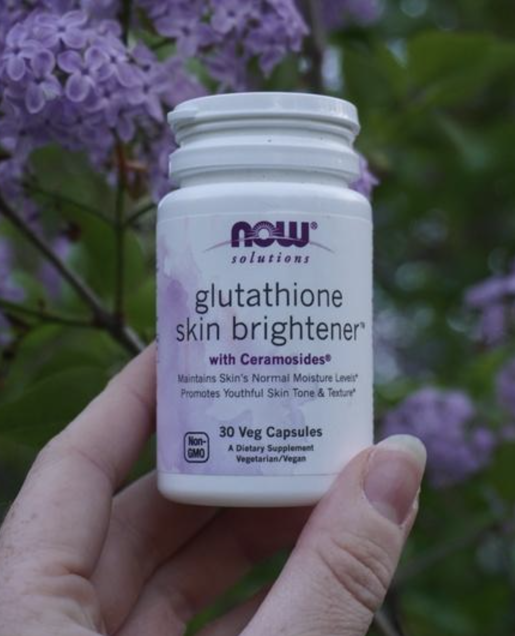 A hand holds a bottle of NOW Solutions Glutathione Skin Brightener capsules in front of purple flowering plants.