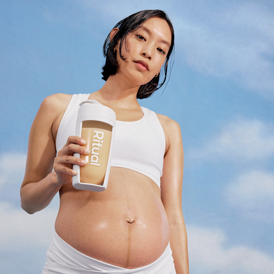 A pregnant person in white athletic wear holds a Ritual-branded bottle against a blue sky background.