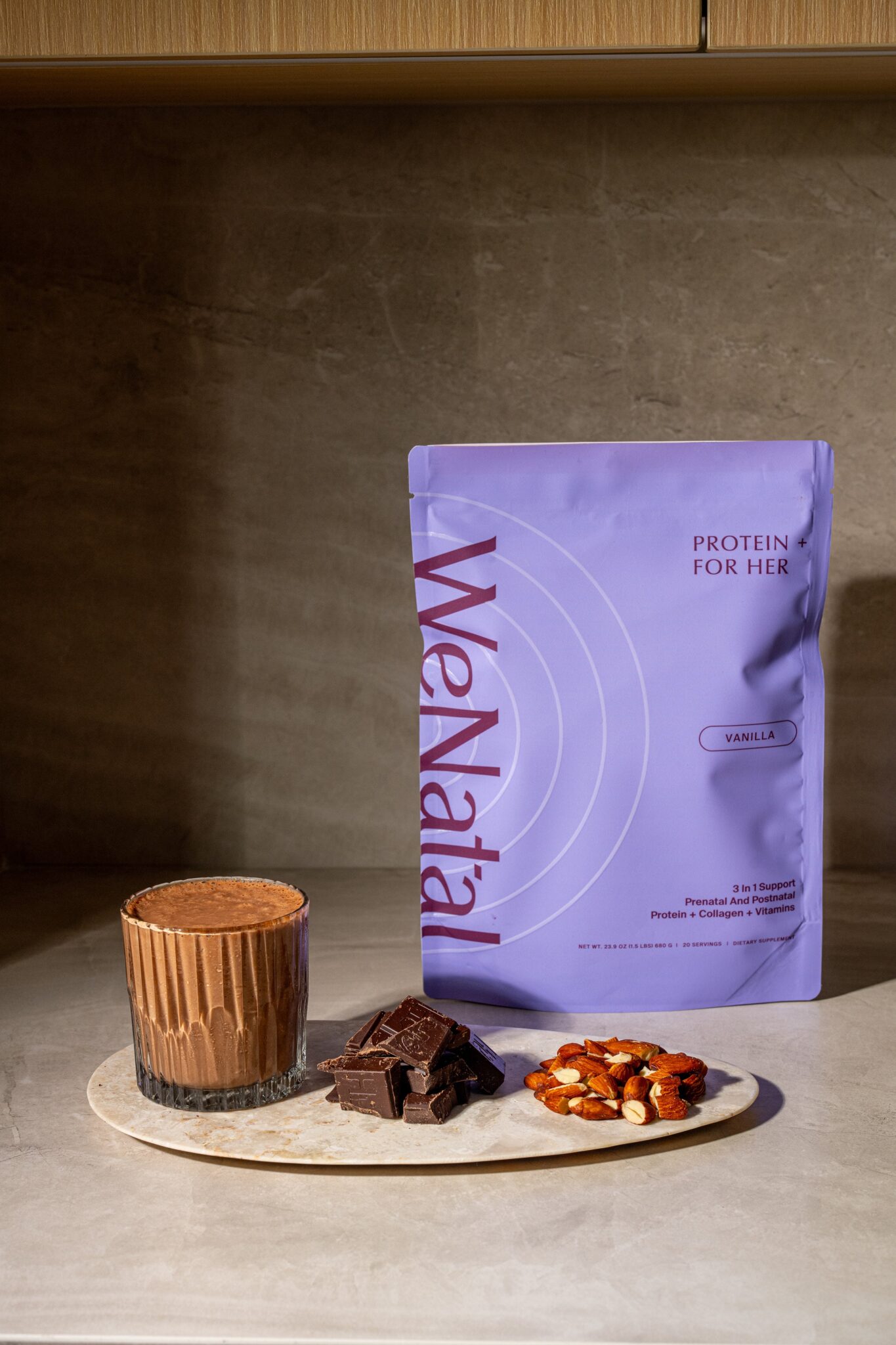 A glass of chocolate protein shake, pieces of chocolate, and mixed nuts are displayed on a plate next to a purple WeNatal Protein For Her vanilla pouch.