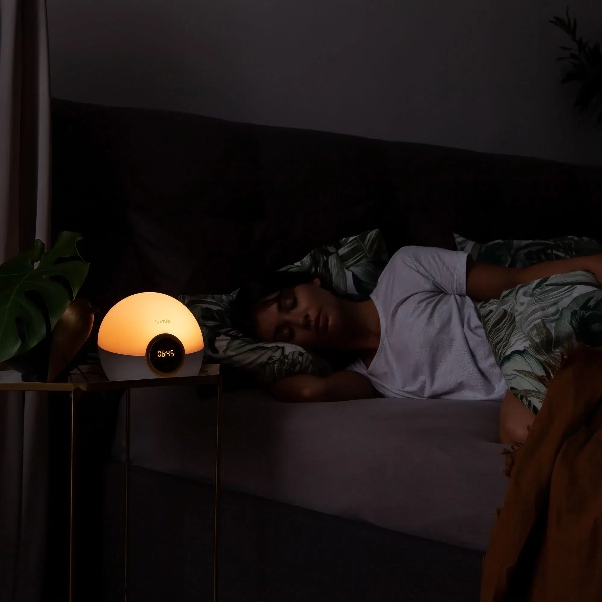 A person sleeps in bed under a patterned blanket while a digital alarm clock with a glowing light sits on a bedside table.