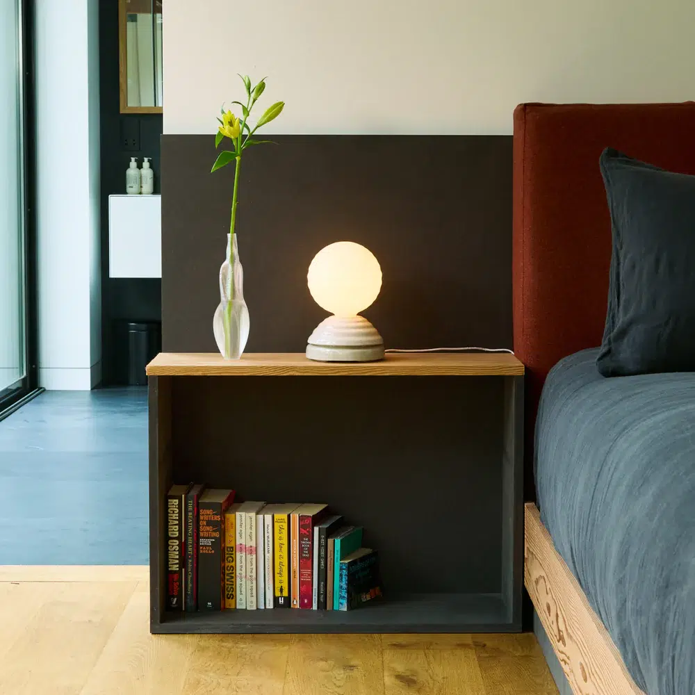 A bedside table with a round lamp, a vase holding a single flower, and a shelf with neatly arranged books next to a bed with dark bedding.