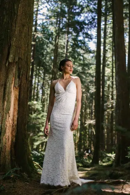 A woman in a white lace dress stands in a sunlit forest, surrounded by tall trees.