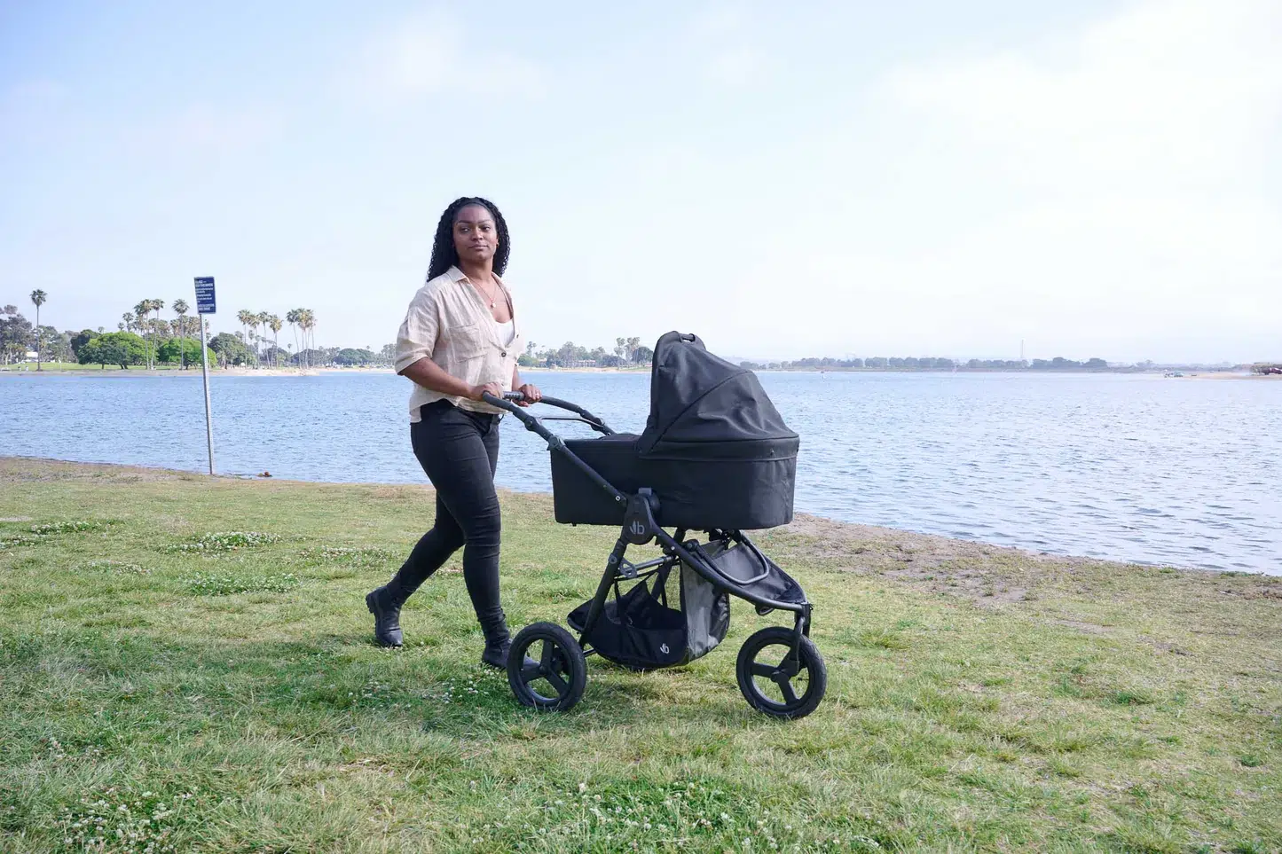 A person pushes a baby stroller along a grassy path beside a body of water on a clear day.