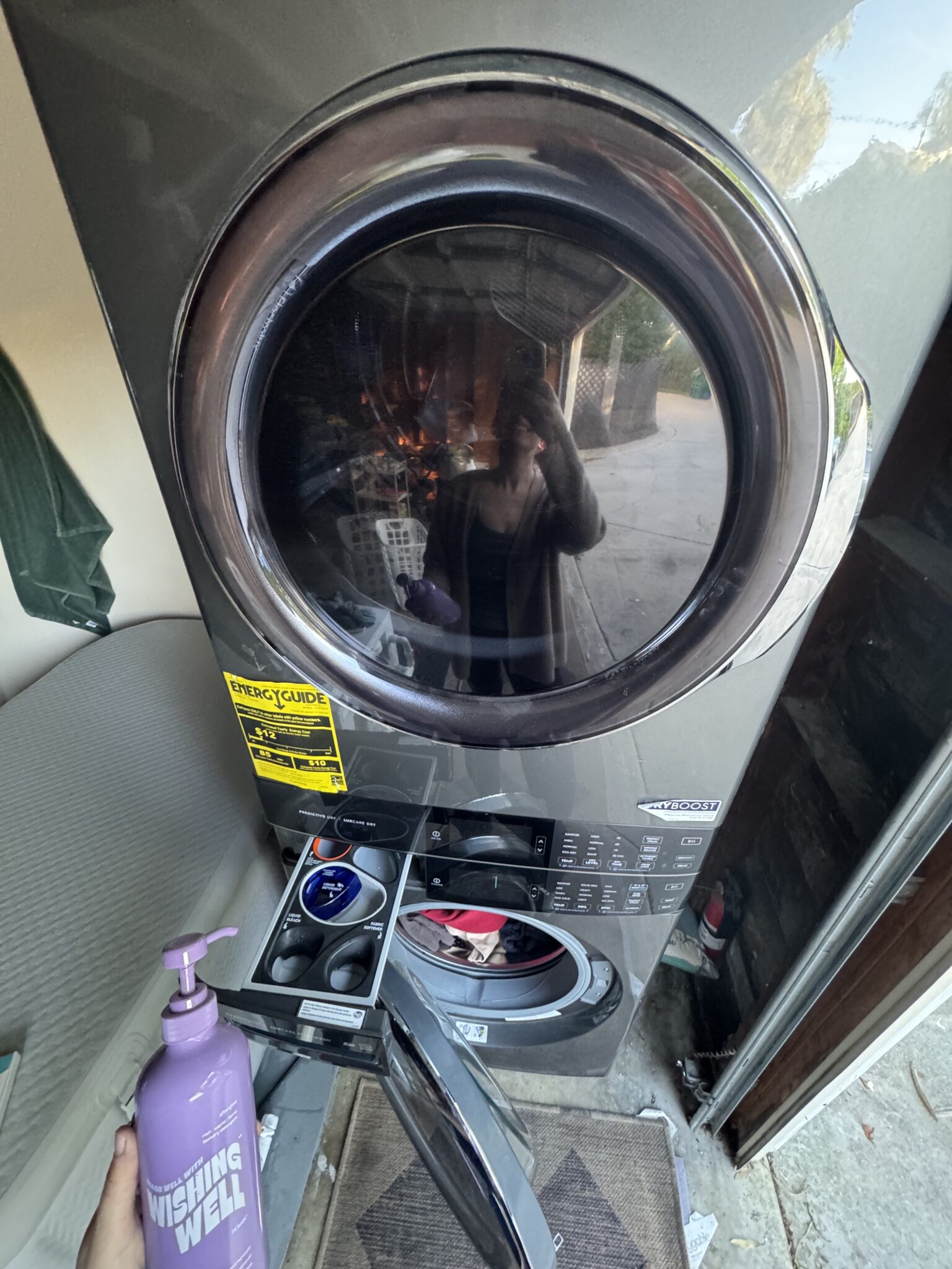 A person holds a bottle of "Wishing Well" soap in front of an open washing machine with detergent tray pulled out, reflecting their image in the door.