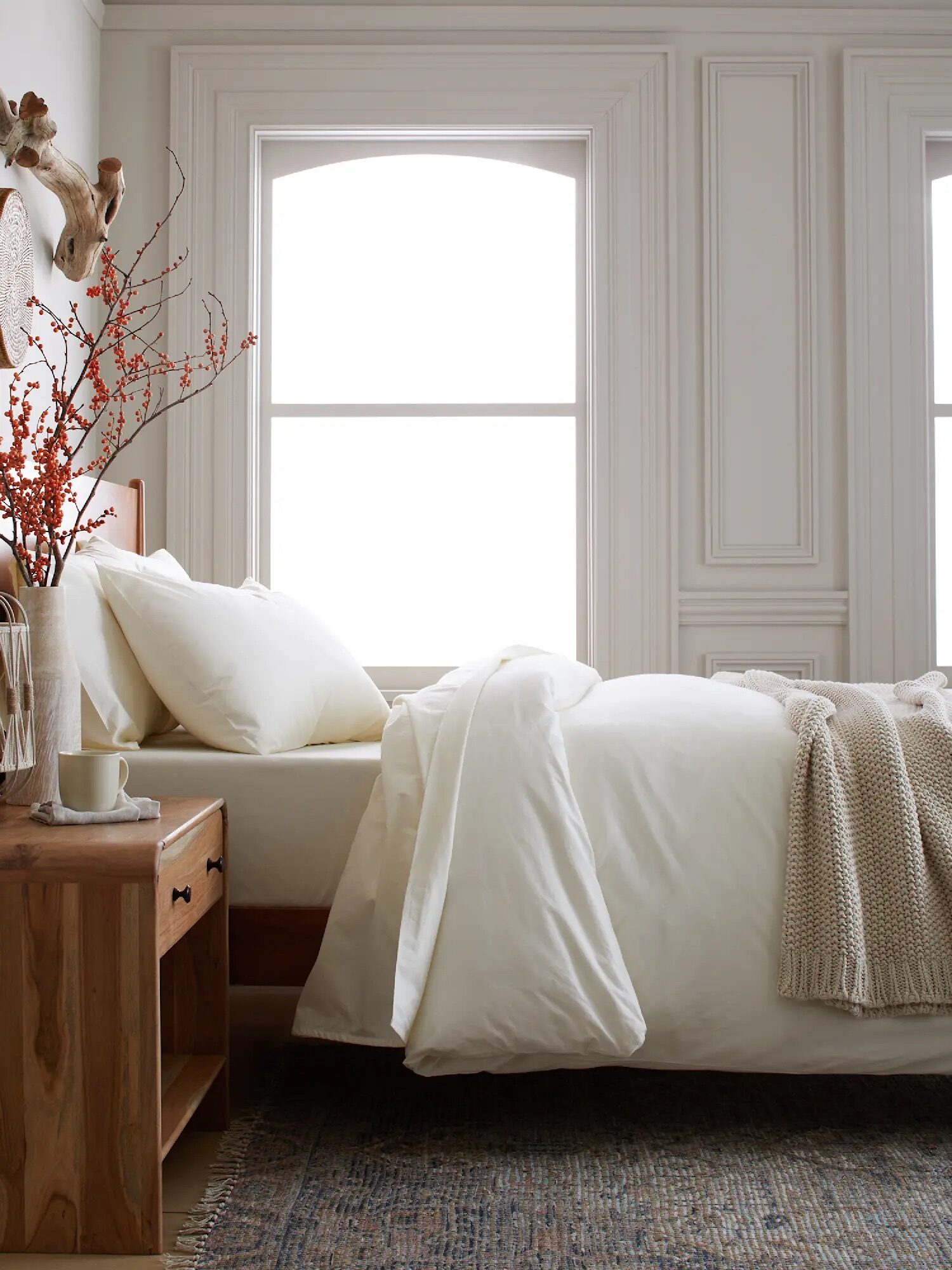 A neatly made bed with white bedding, a beige knit blanket, and a wooden nightstand with a mug and vase of red berries, set in a bright room with two large windows.