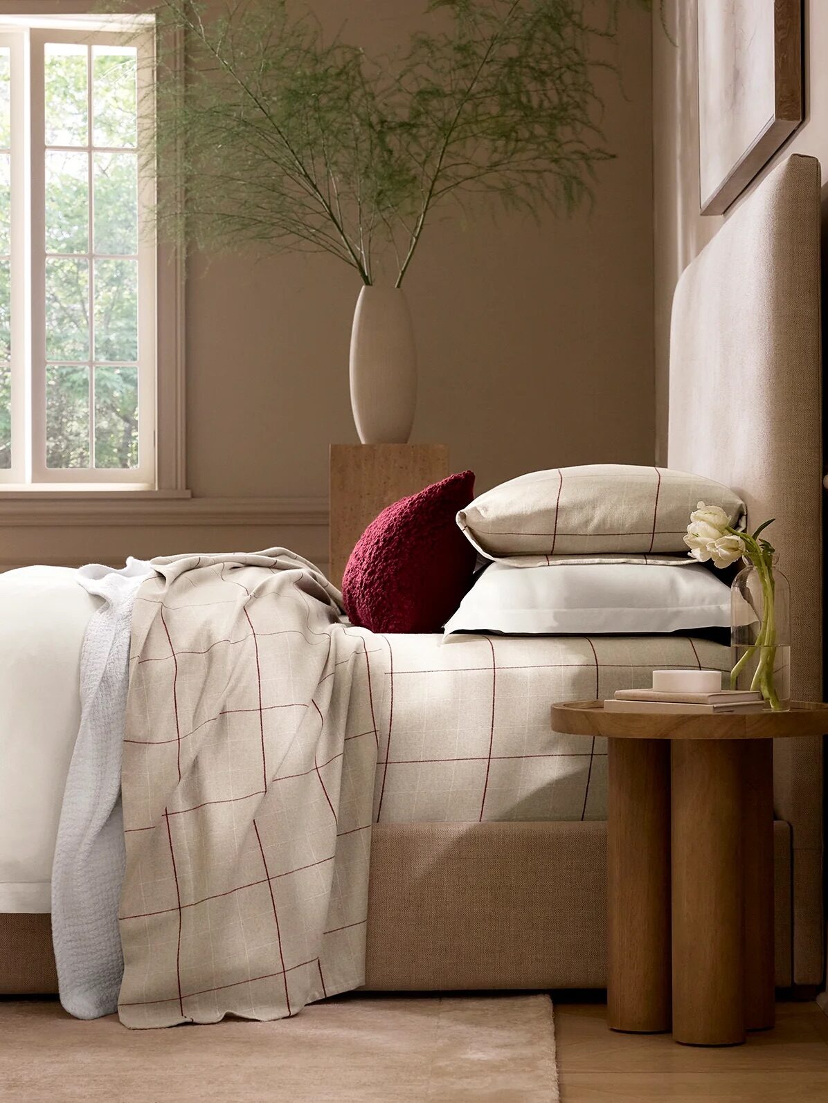 A neatly made bed with neutral checked bedding, a stack of pillows, a red throw, and a small wooden side table holding a flower vase in a softly lit bedroom.