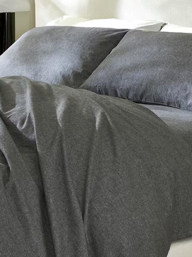 A neatly made bed with dark gray sheets and pillowcases, a slightly rumpled top sheet, and a bedside table with a plant and lamp.