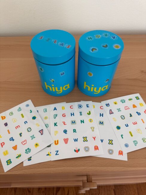 Two blue cylindrical containers labeled "hiya" on a wooden surface, with sheets of colorful alphabet stickers spread out in front of them.