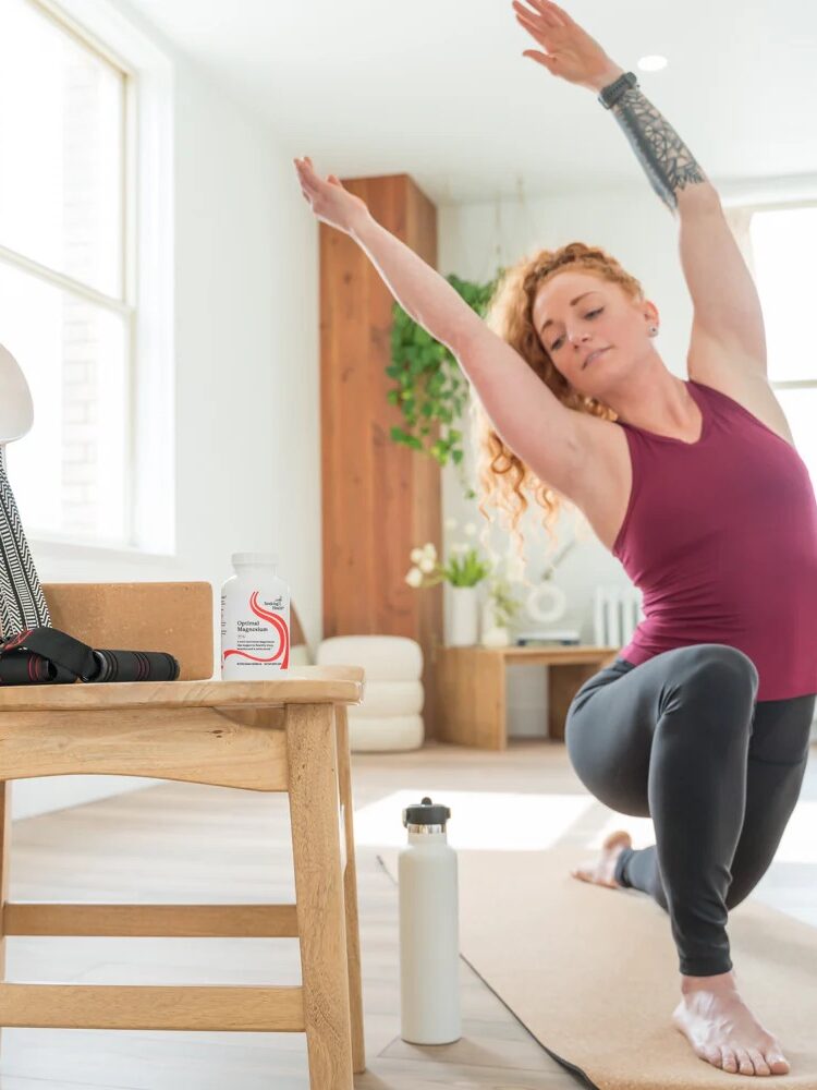 A woman practices a side-stretch yoga pose on a mat in a bright room, with a water bottle, supplements, and resistance bands on a nearby chair.