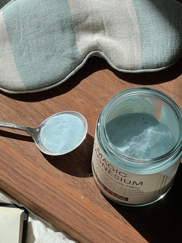 A spoon with blue magnesium powder rests next to an open jar labeled "Magnesium" on a wooden tray, with a striped sleep mask and part of an open book nearby.