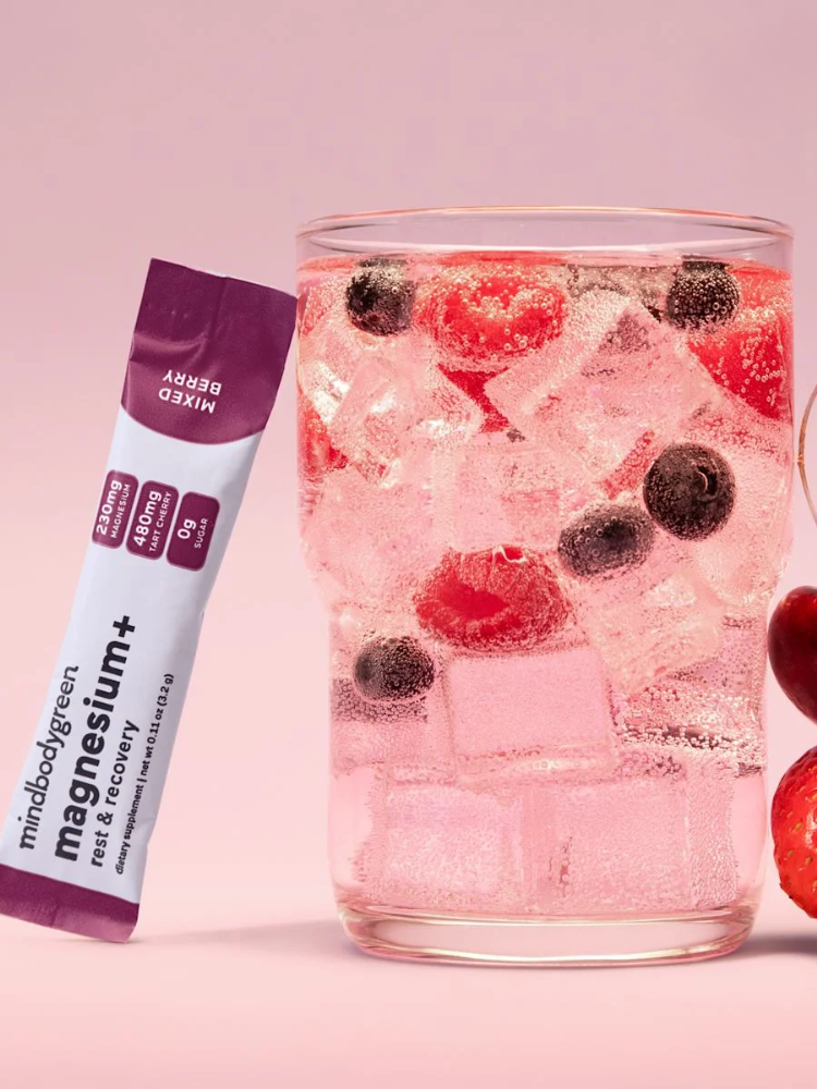 A glass of sparkling water with ice, strawberries, and blueberries sits next to a packet labeled "mindbodygreen magnesium+ mixed berry.