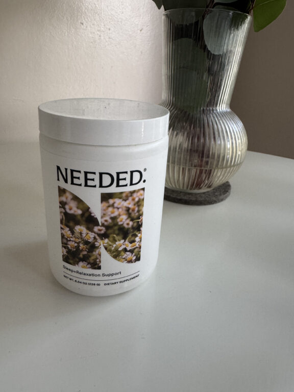 A white container labeled “NEEDED: Sleep+Relaxation Support” sits on a white surface near a ribbed glass vase with green leaves.