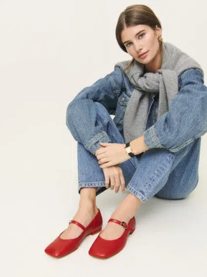 A woman wearing blue jeans, a denim jacket, a gray scarf, and red Mary Jane shoes sits on the floor against a plain background.