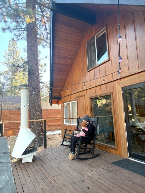 A person sits on a wooden deck outside a cabin, holding a baby on their lap in a rocking chair. A white outdoor fireplace and tall pine tree are nearby.