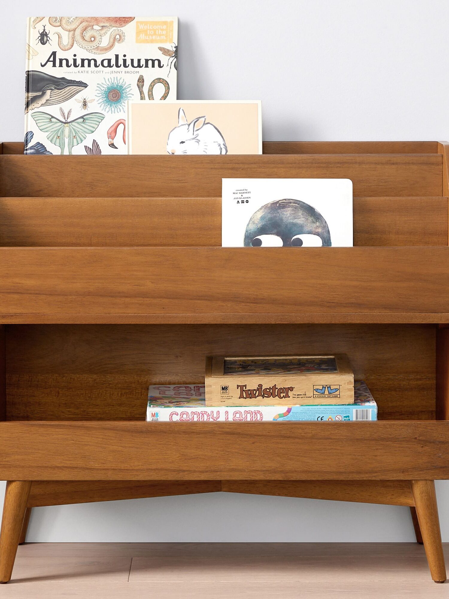 A wooden bookshelf with three shelves holds children's books, including "Animalium," and board games on the bottom shelf, against a light-colored wall.