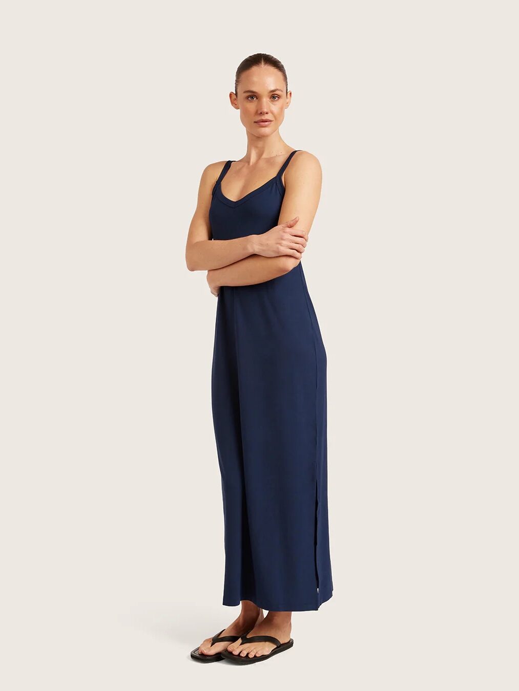 A woman stands with arms crossed, wearing a navy blue sleeveless maxi dress and black sandals against a plain, light background.