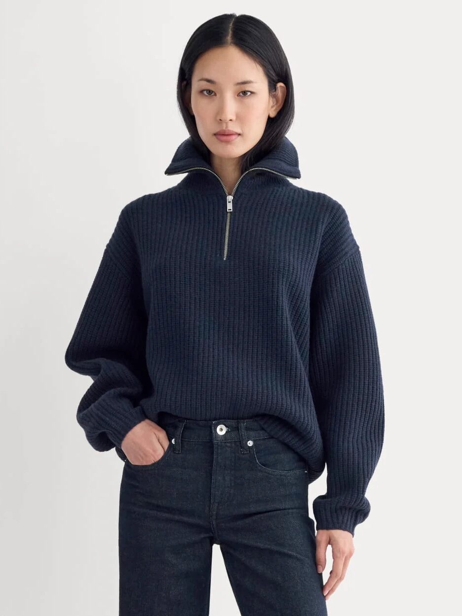 A woman wearing a dark blue ribbed half-zip pullover sweater and black jeans stands against a plain light background.
