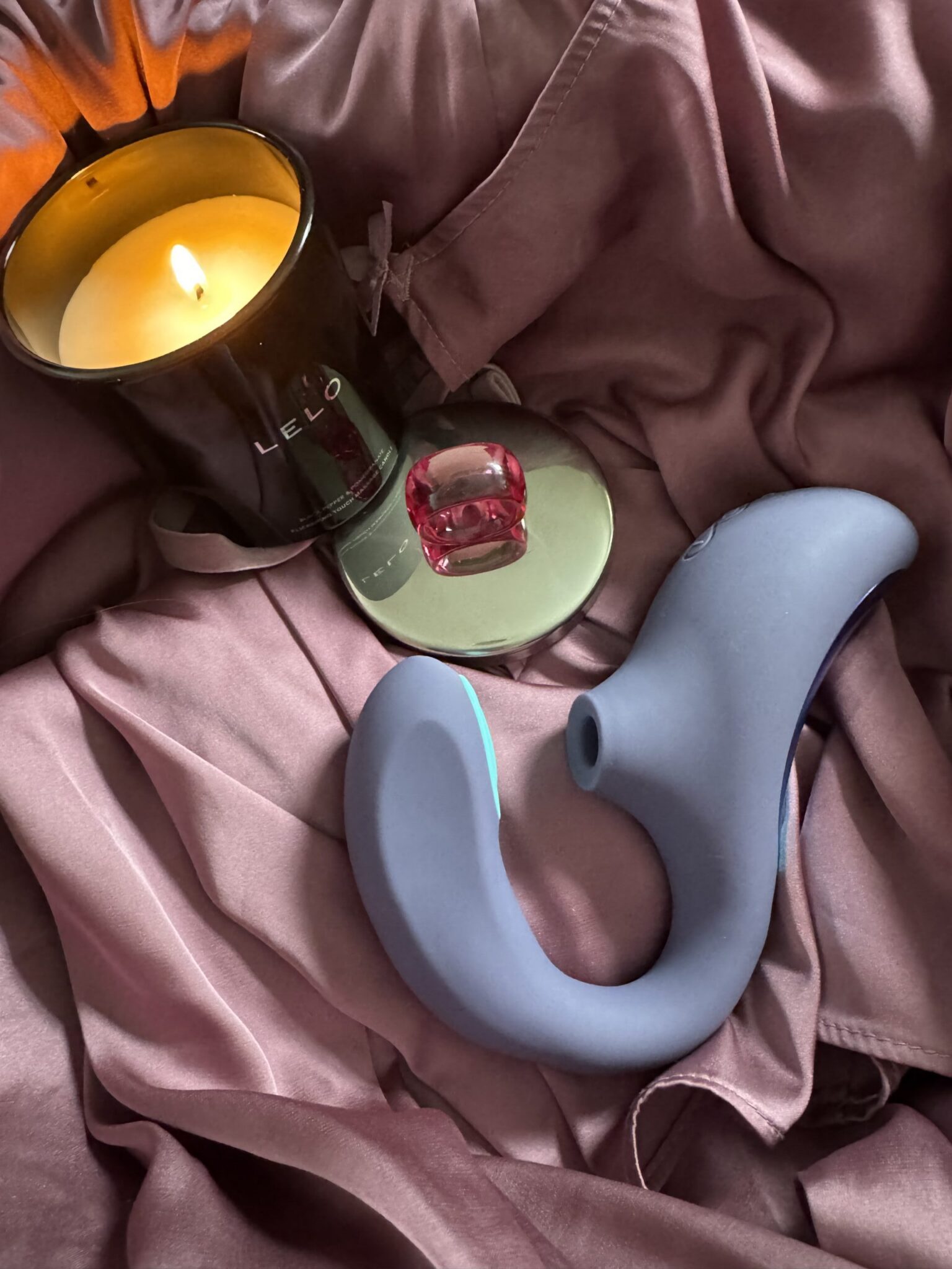 A lit candle, a red glass ring, and a blue curved sex toy are arranged on mauve satin fabric.