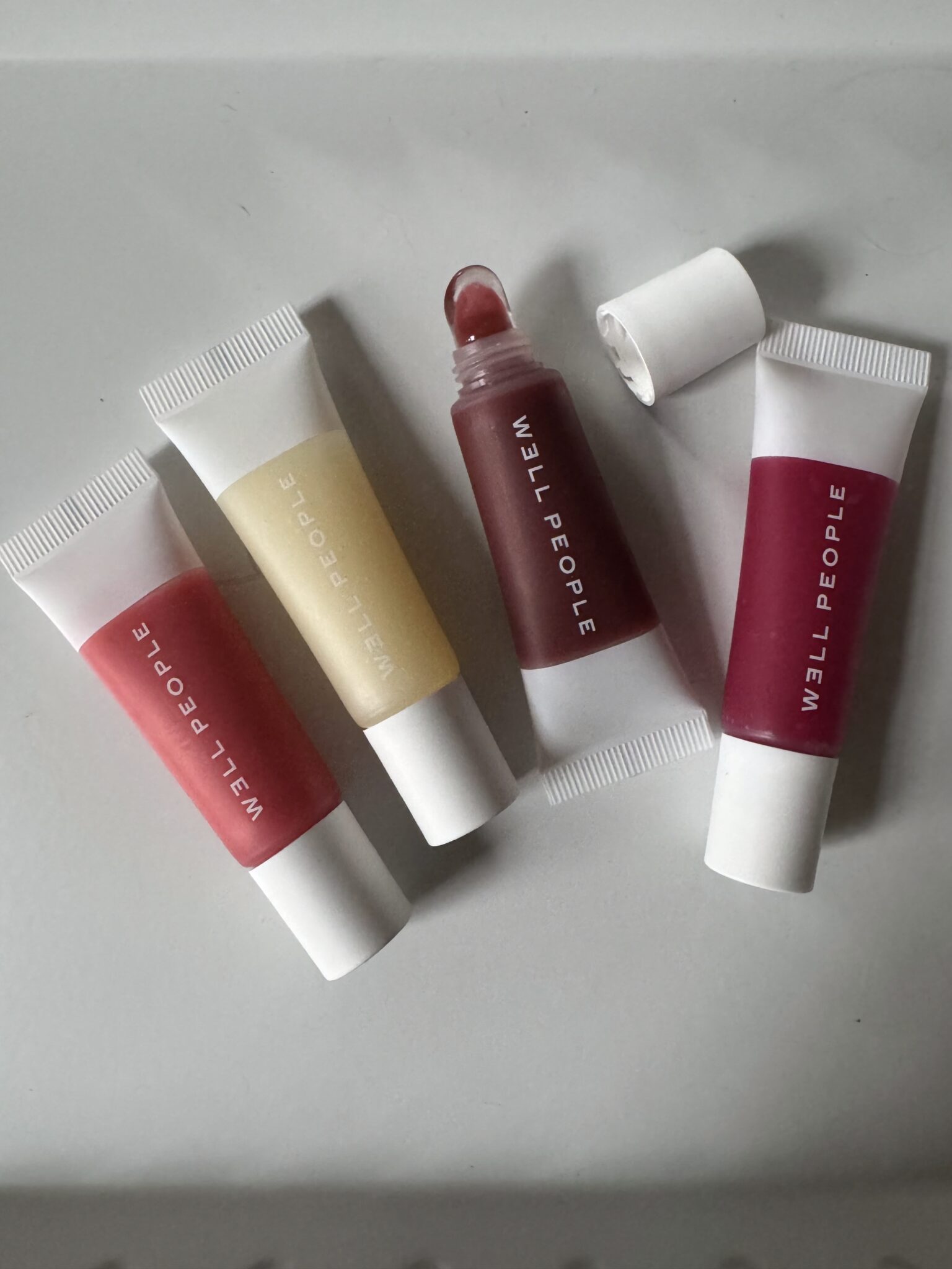Four tubes of WELL PEOPLE lip balm in different shades are arranged on a flat surface, with one tube open and its cap placed nearby.