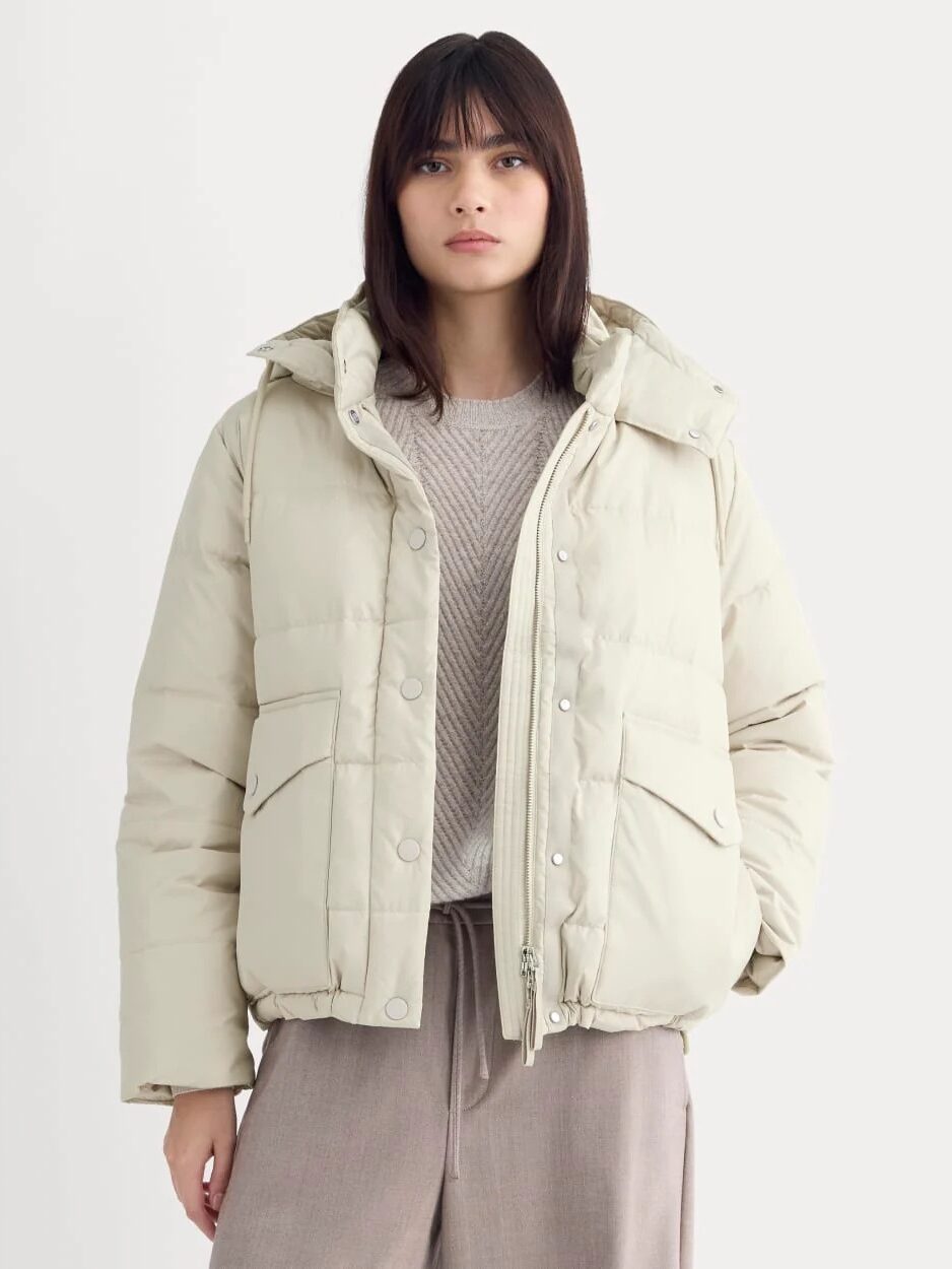 A person wearing a light beige puffer jacket over a textured sweater and loose tan pants, standing against a plain light background.