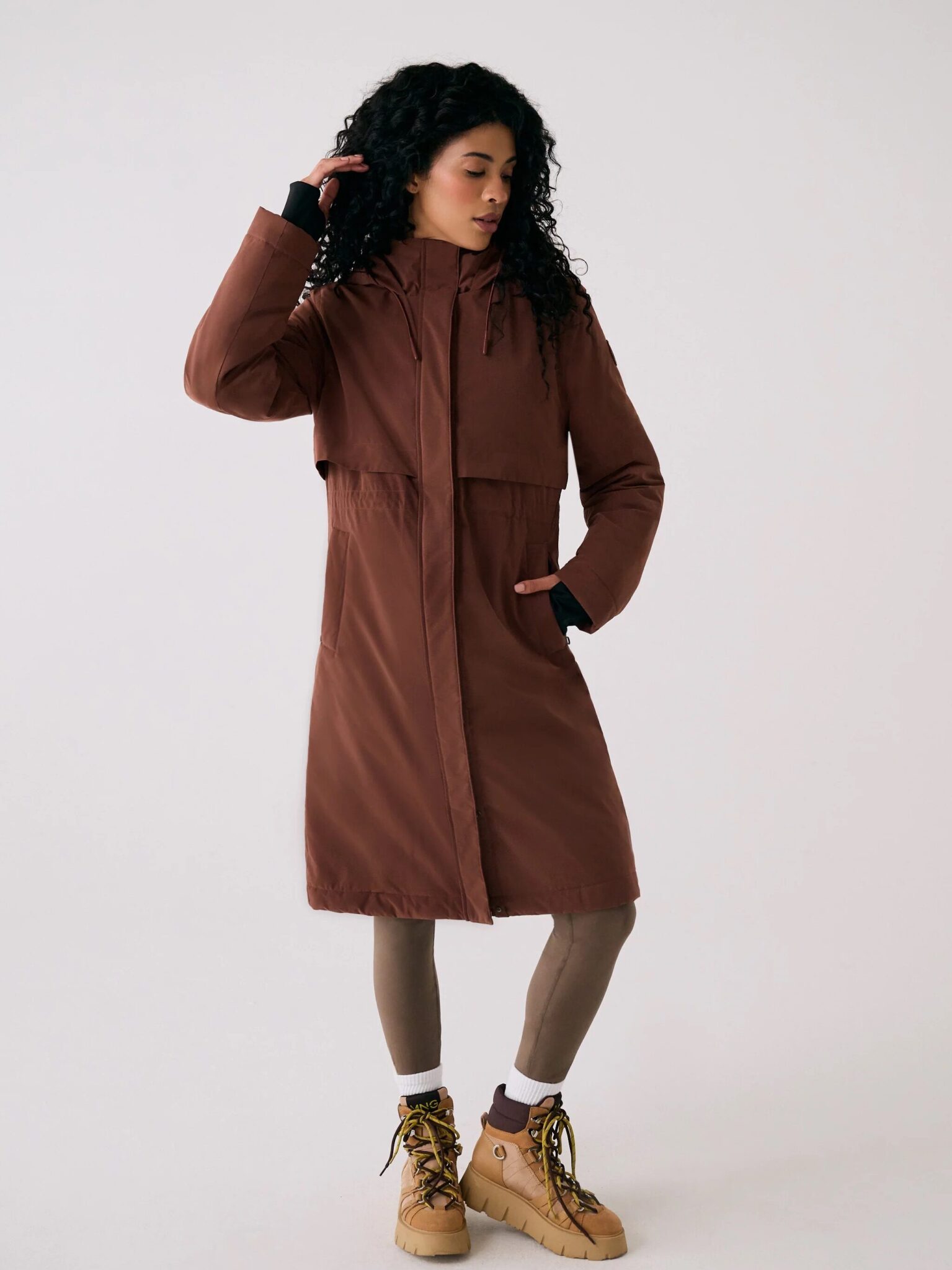 A person wearing a long brown hooded coat, taupe leggings, and tan lace-up boots stands against a plain white background.