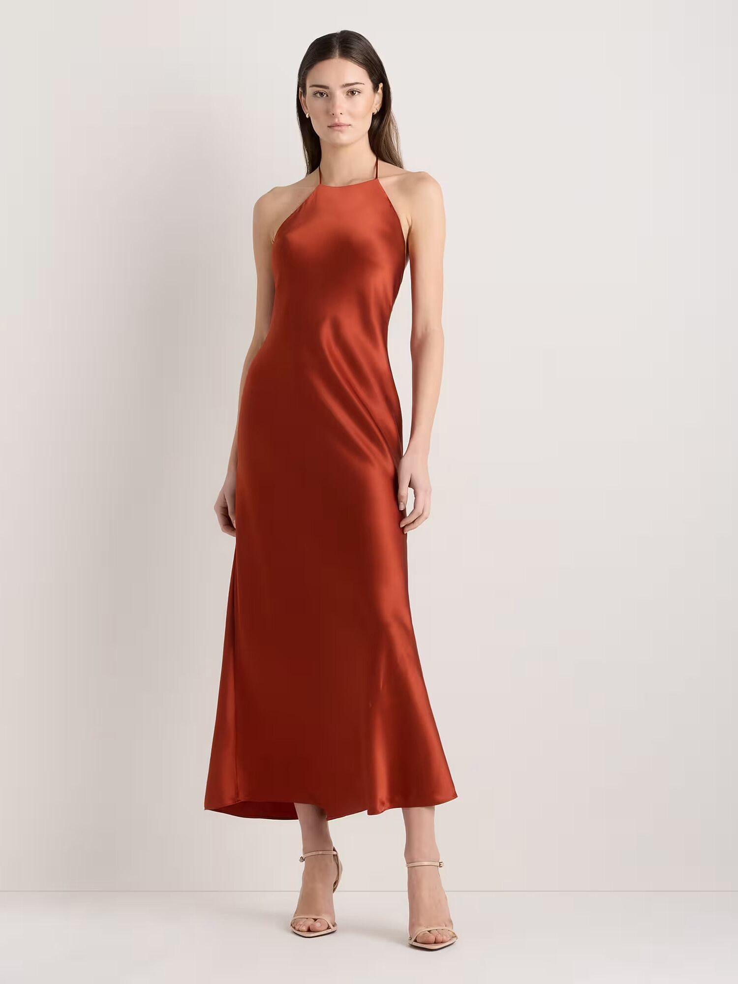 A woman stands against a plain background wearing a sleeveless, rust-colored satin halter dress and nude heels.