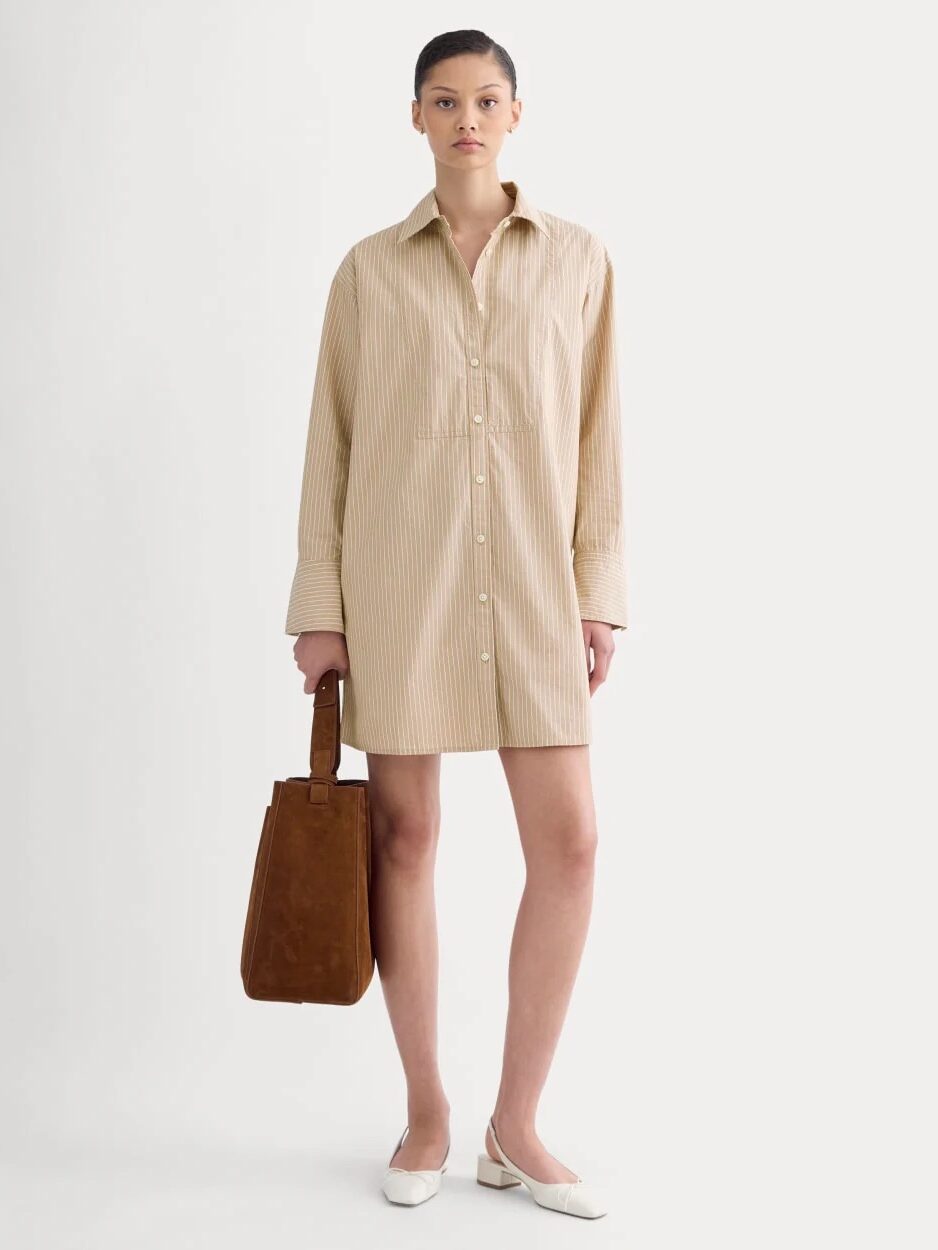 A woman stands against a plain background wearing a beige striped shirt dress, holding a brown suede tote bag, and white flats.