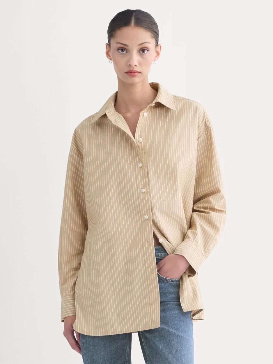A woman wearing a beige striped button-up shirt and blue jeans stands against a plain light background, looking at the camera with a neutral expression.