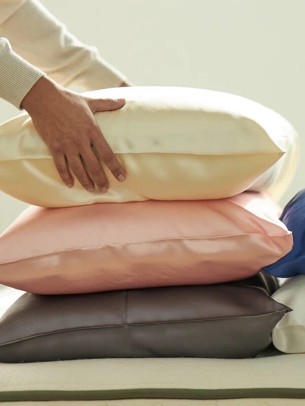 A person arranging a stack of four satin pillowcases in cream, pink, gray, and blue colors on a surface.