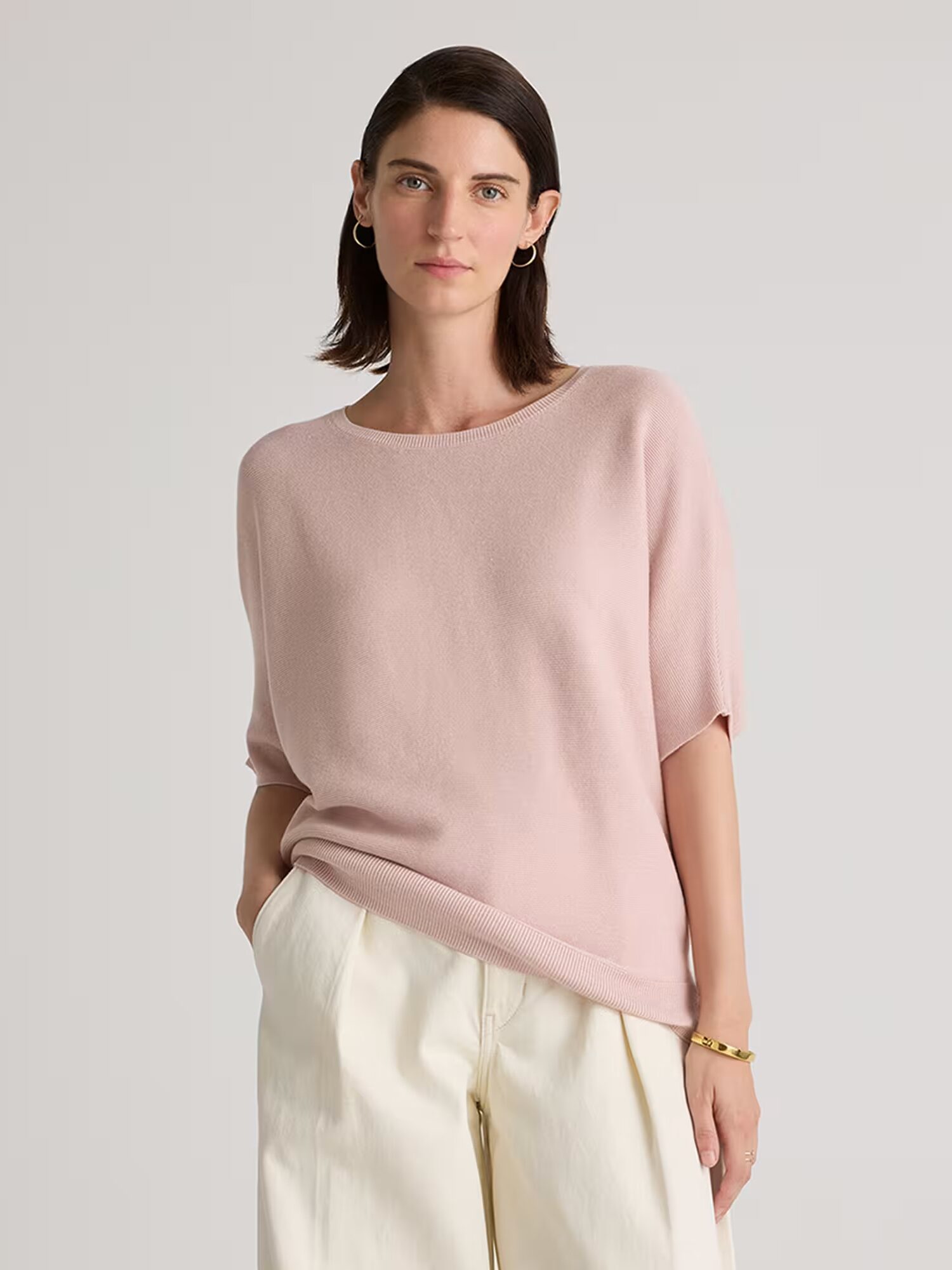 Woman wearing a light pink short-sleeve sweater and cream pants stands with one hand in her pocket against a neutral background.