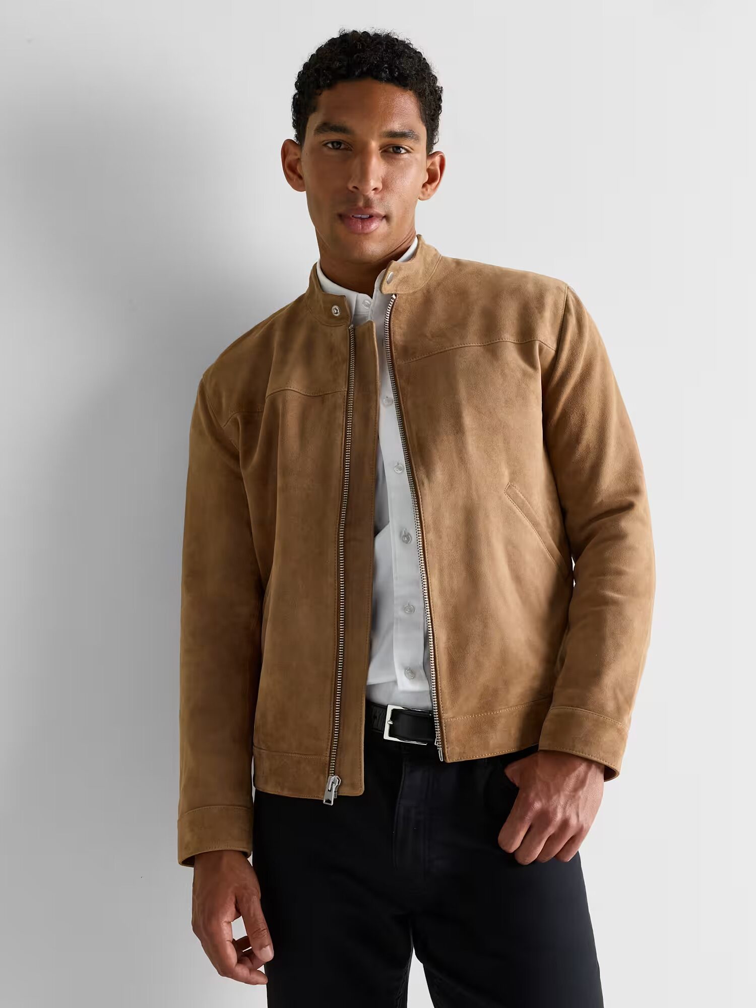 A man wearing a tan suede jacket over a white button-up shirt and black pants stands against a plain light gray background.
