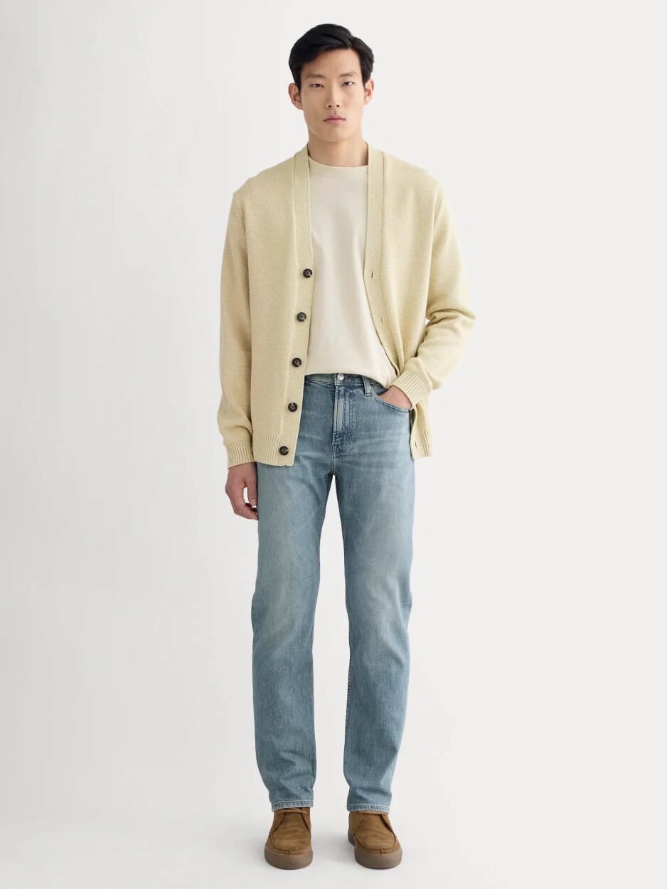 A person stands facing forward wearing a cream cardigan, white t-shirt, light blue jeans, and brown shoes against a plain light background.