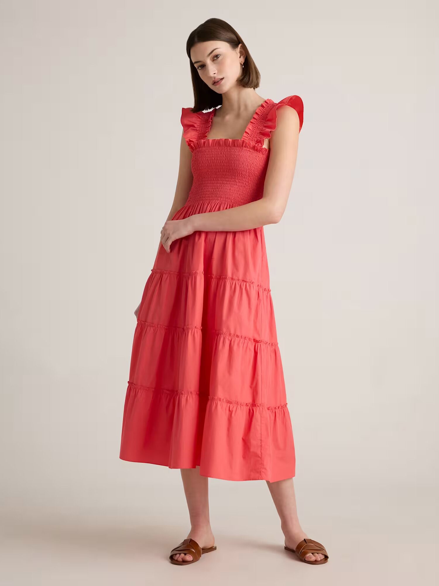 A woman stands wearing a coral pink, tiered midi dress with ruffled sleeves and brown flat sandals against a plain background.