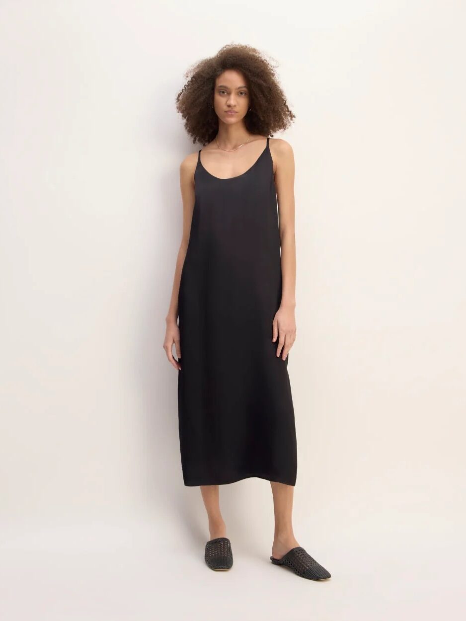 A woman stands against a plain background wearing a sleeveless, loose-fitting black midi dress and black slip-on shoes.