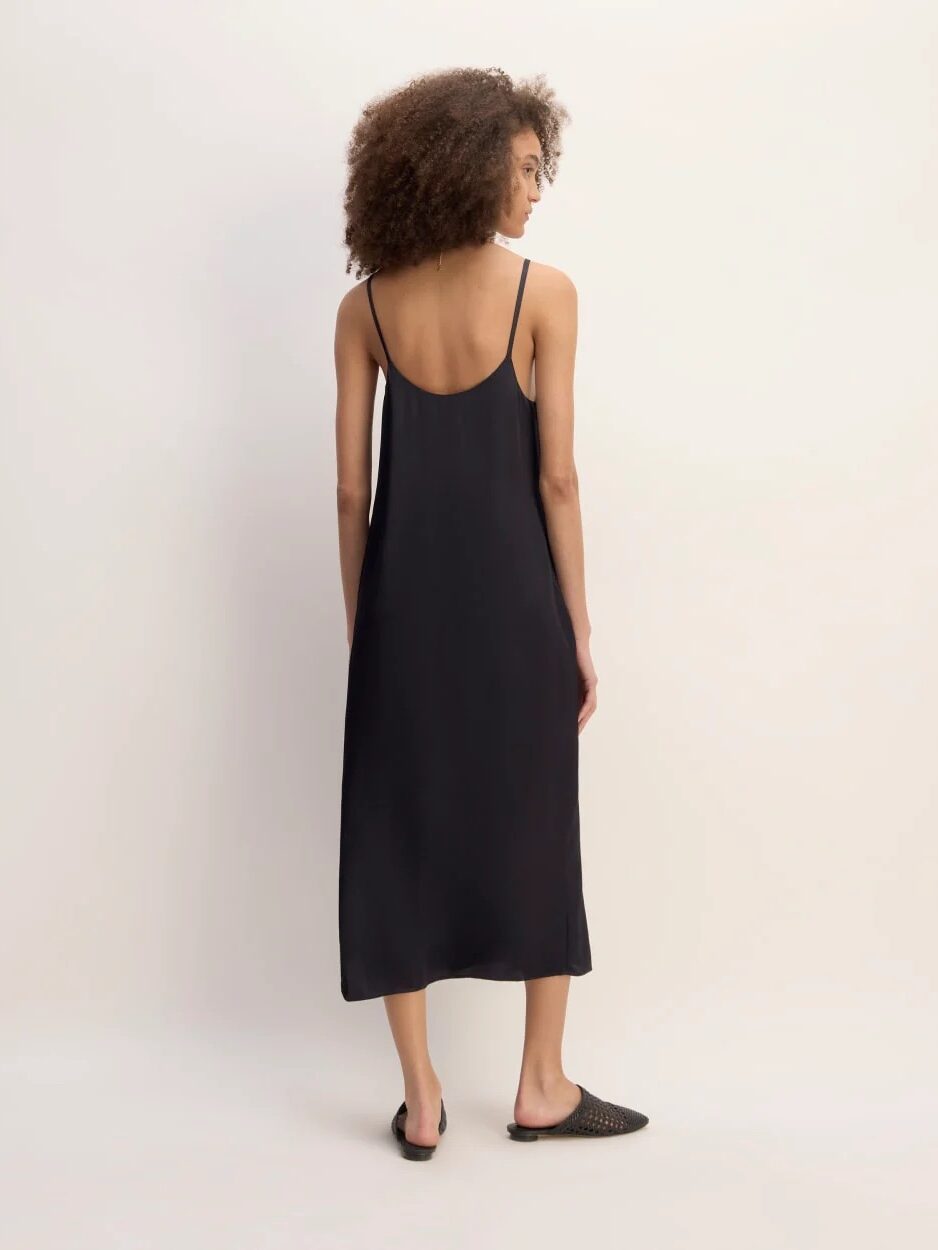 A woman stands facing away from the camera, wearing a black sleeveless midi slip dress with thin straps and black slip-on shoes against a plain light background.