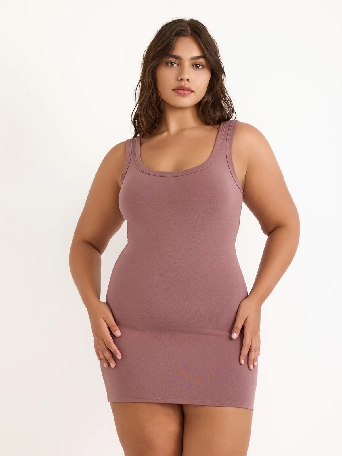 Woman standing against a plain background wearing a fitted, sleeveless, mauve mini dress.