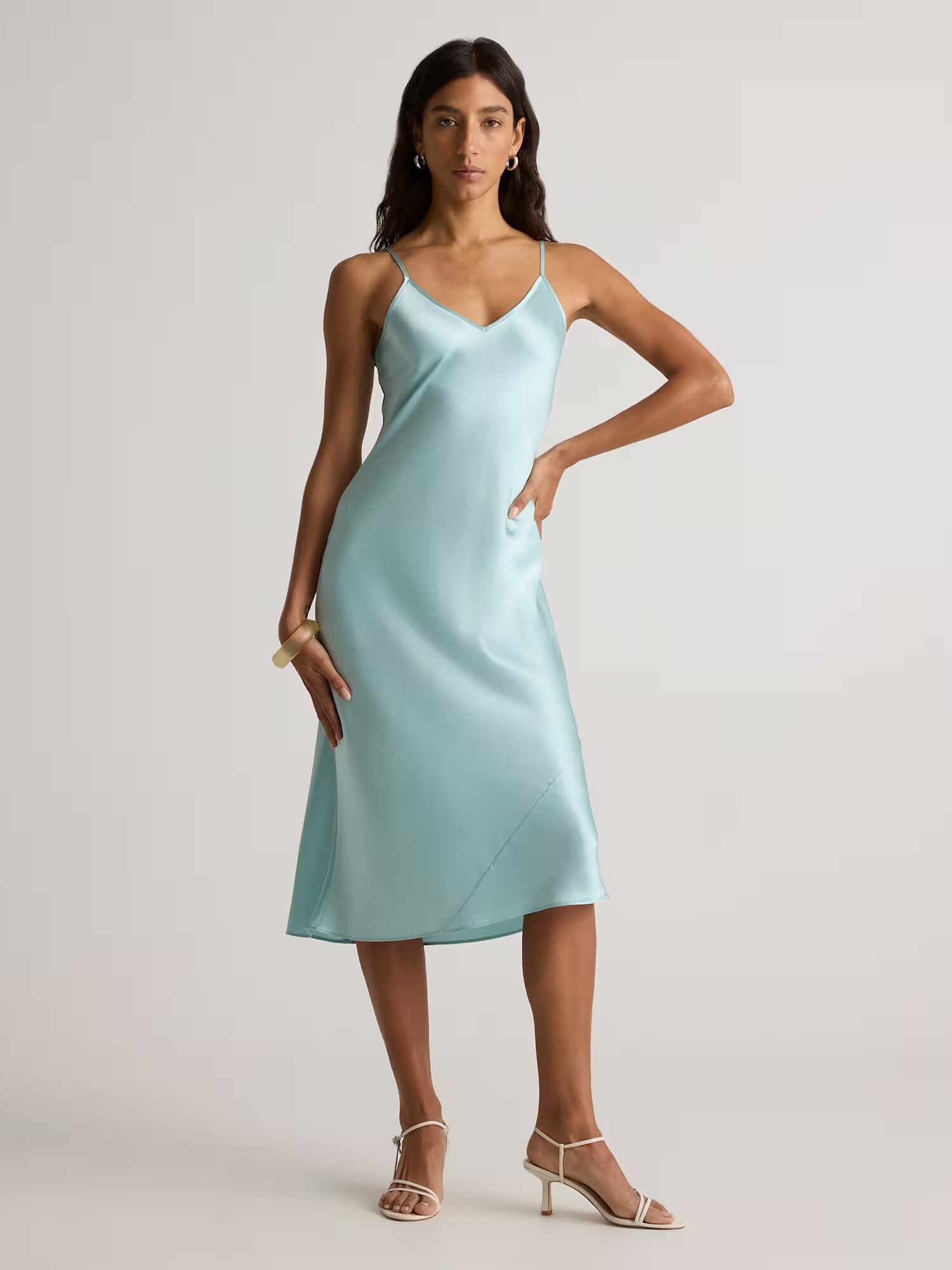 Woman wearing a light blue, sleeveless, midi-length slip dress with thin straps and beige heeled sandals, standing against a neutral background.