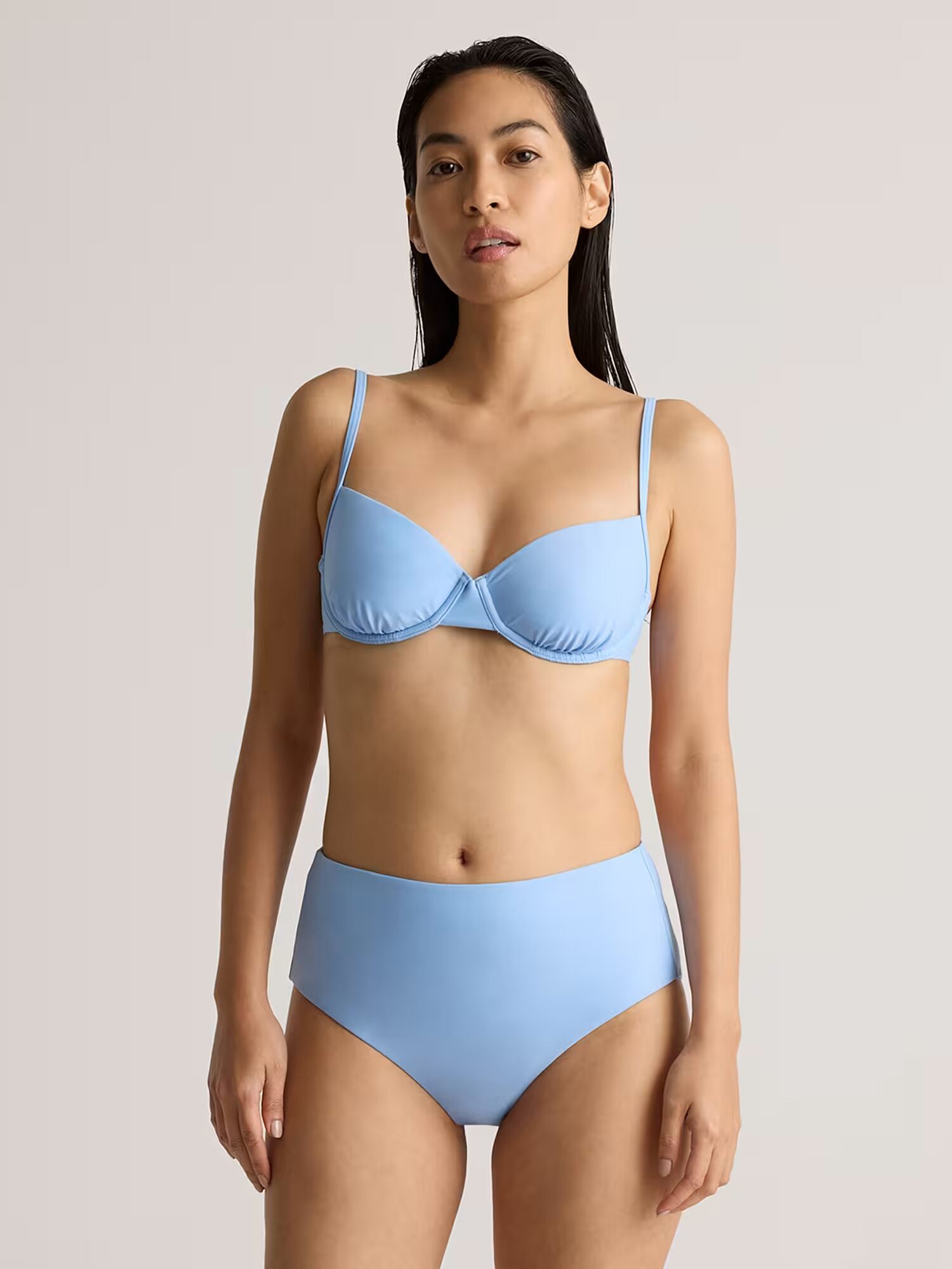 A woman stands facing forward, wearing a light blue bikini top and high-waisted bikini bottoms against a plain background.