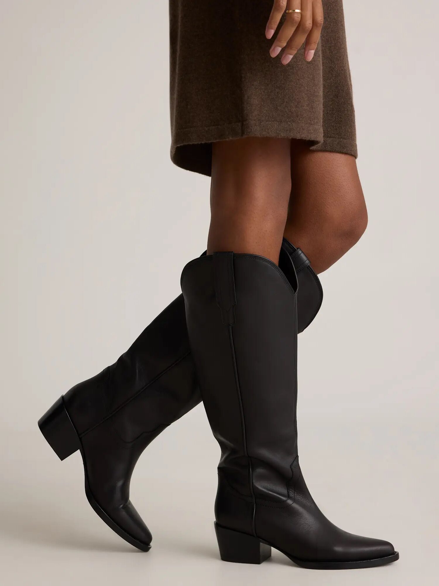 A person wearing knee-high black leather boots and a brown skirt stands on a neutral background.