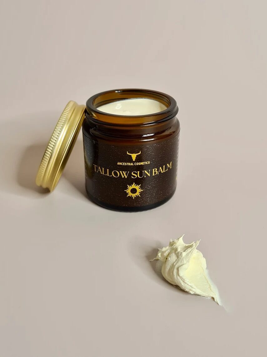 An open brown jar labeled "Tallow Sun Balm" with a gold lid and a dollop of cream on a neutral surface.