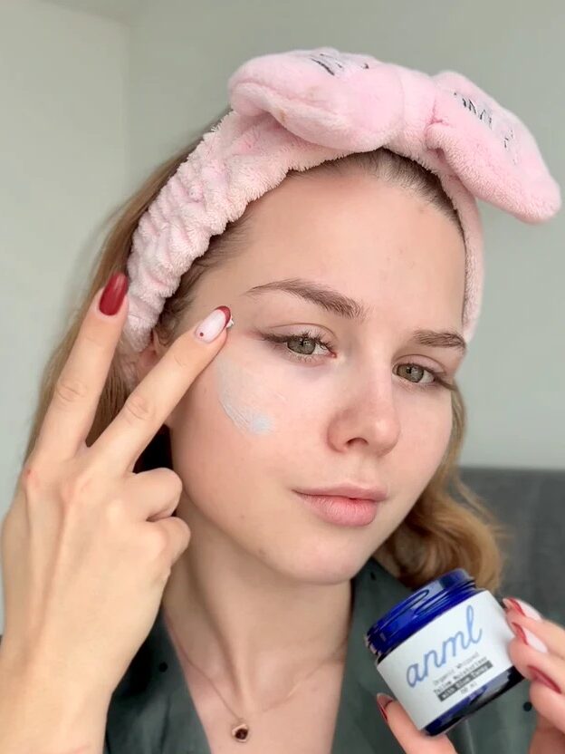 A woman wearing a pink headband applies cream to her cheek with her fingers while holding a blue jar labeled "anml.
