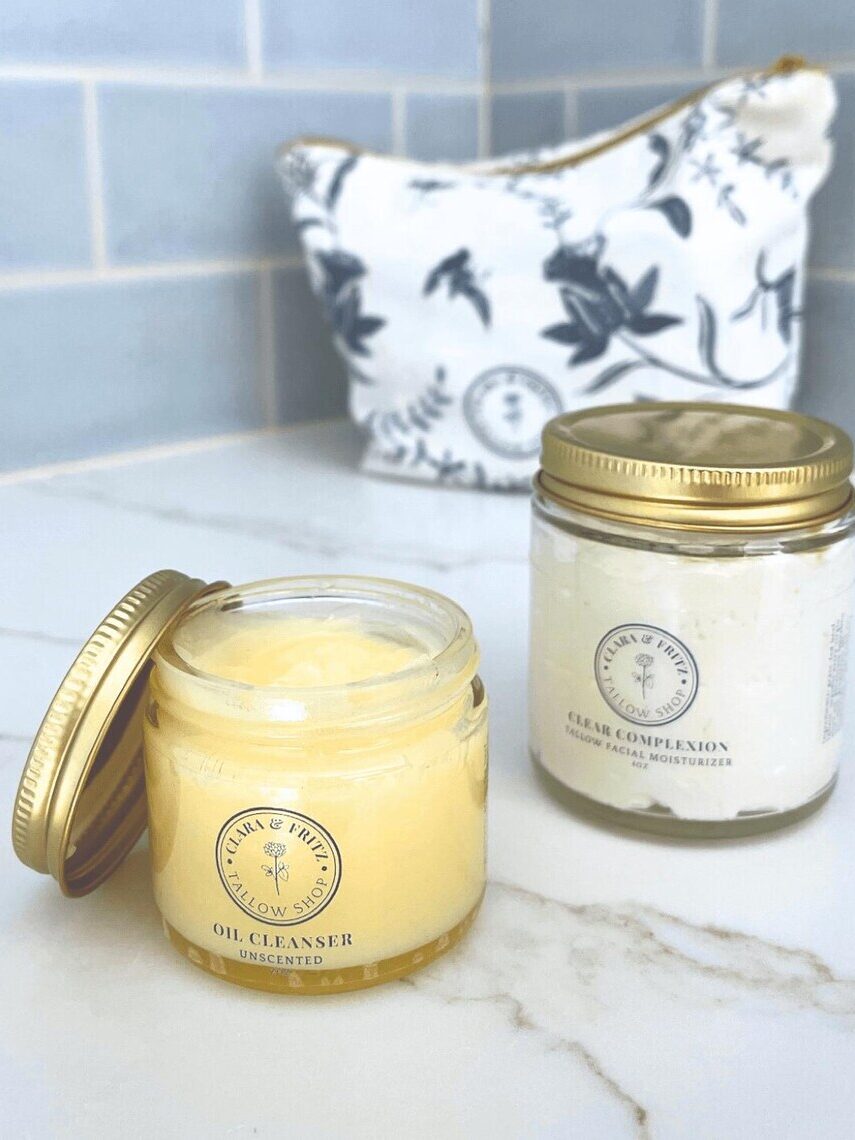 Two jars of skincare products, one labeled "Oil Cleanser" with its lid off and one labeled "Facial Moisturizer," sit on a marble surface with a patterned pouch in the background.
