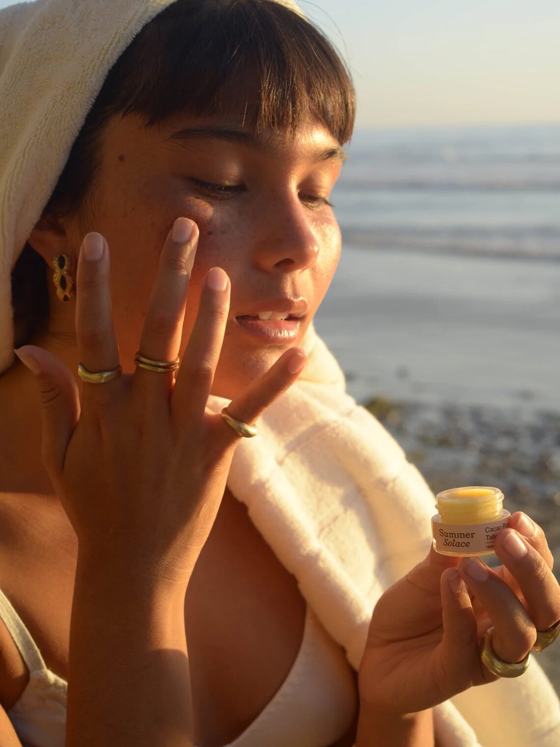 A person with a towel on their head applies a cream to their face while holding a small jar labeled "Summer Solace" by the beach.