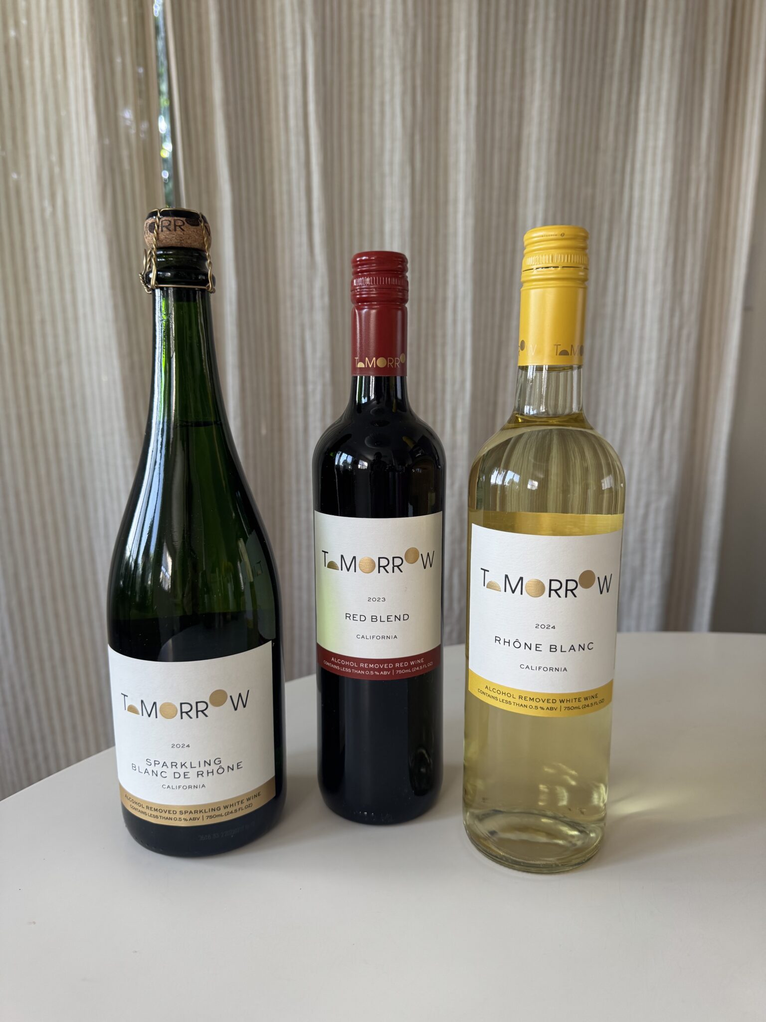 Three bottles of Tomorrow brand wine—sparkling blanc de noir, red blend, and 2022 Rhone blanc—stand on a white table in front of beige curtains.